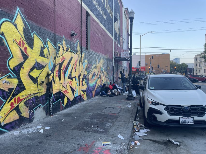 People gather on a littered sidewalk beside a building with large, colorful graffiti; a white car is parked in the foreground.