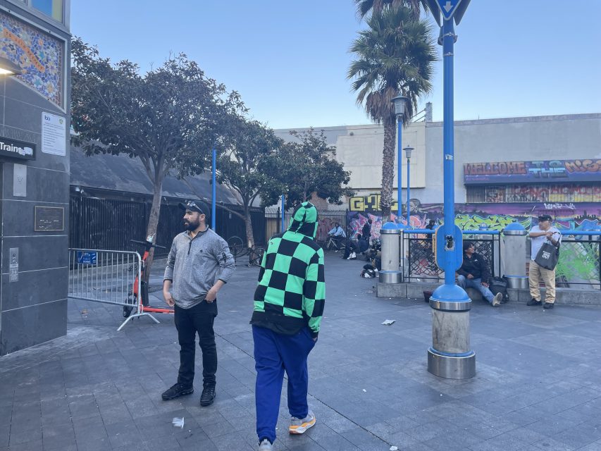 A city street scene with several people standing and sitting near colorful graffiti walls, trees, and a palm tree; scattered litter is visible on the ground.