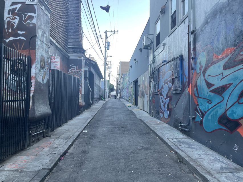 A narrow urban alleyway with graffiti-covered walls, power lines overhead, and a pair of shoes hanging from a wire.