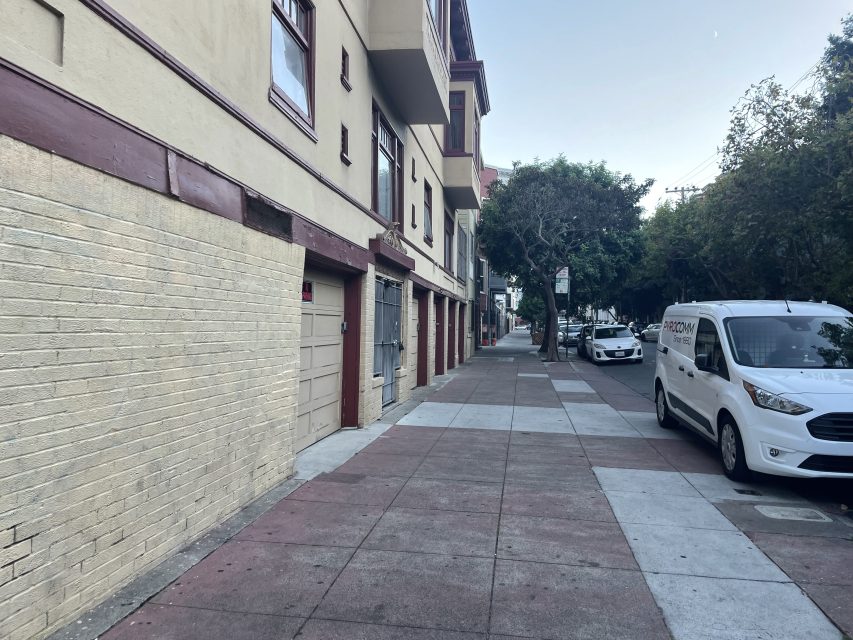 A city sidewalk lined with trees, parked cars, and beige buildings with maroon trim on a clear day.