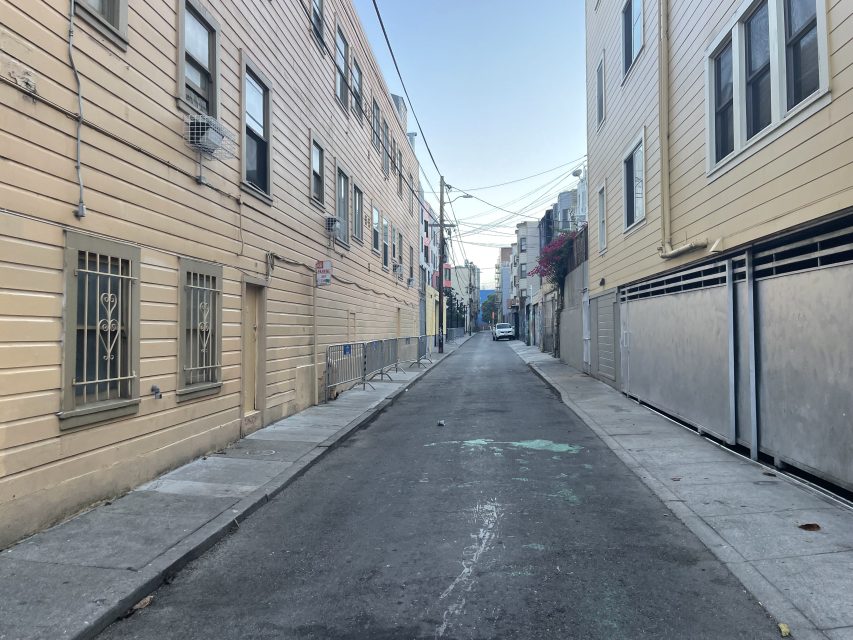 A narrow urban alleyway lined with beige buildings on both sides, with metal barriers on the left and a single car parked at the far end under a clear sky.