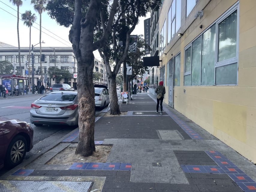 A city sidewalk with trees, parked cars, and a person wearing a red hat walking past a yellow building with large windows.