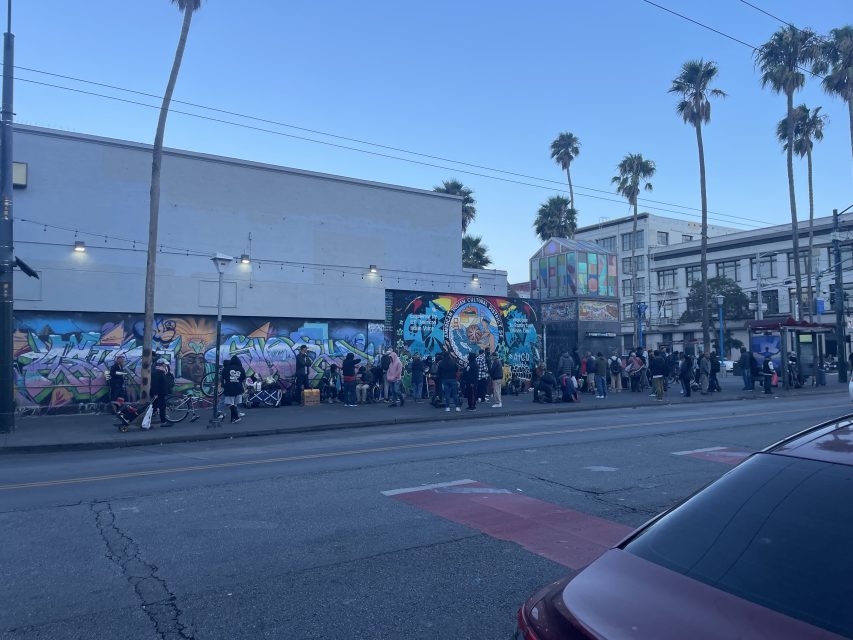 A group of people stand in line on a city sidewalk in front of a mural-covered wall, with palm trees and buildings in the background.