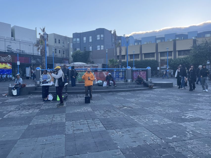 A group of people gather in an urban square with musicians performing and others sitting or walking nearby; buildings and murals are visible in the background.