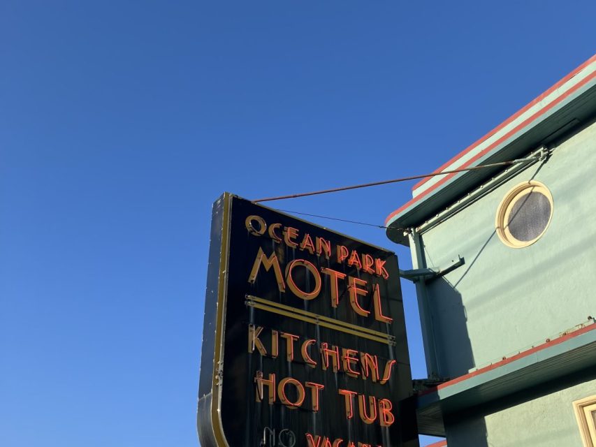 A neon sign for Ocean Park Motel advertises kitchens and a hot tub, mounted on a green building with a round window, against a clear blue sky.