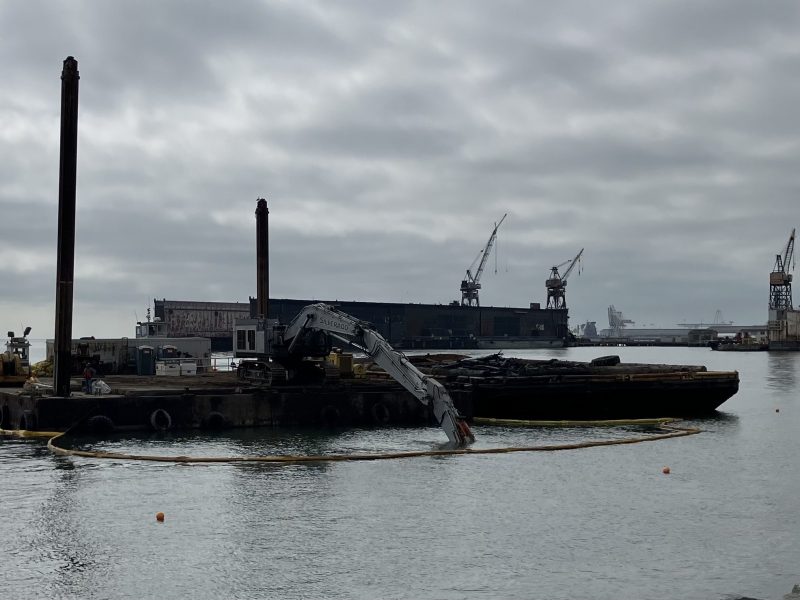 A barge with a long-arm excavator is positioned in the water near a harbor, surrounded by cranes and industrial structures under a cloudy sky.