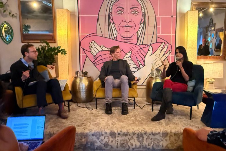 Three people sit and converse on a small stage in a cozy room, discussing harm reduction, with a large mural of a woman and the word "TOGETHER" behind them. Audience members are visible in the foreground.