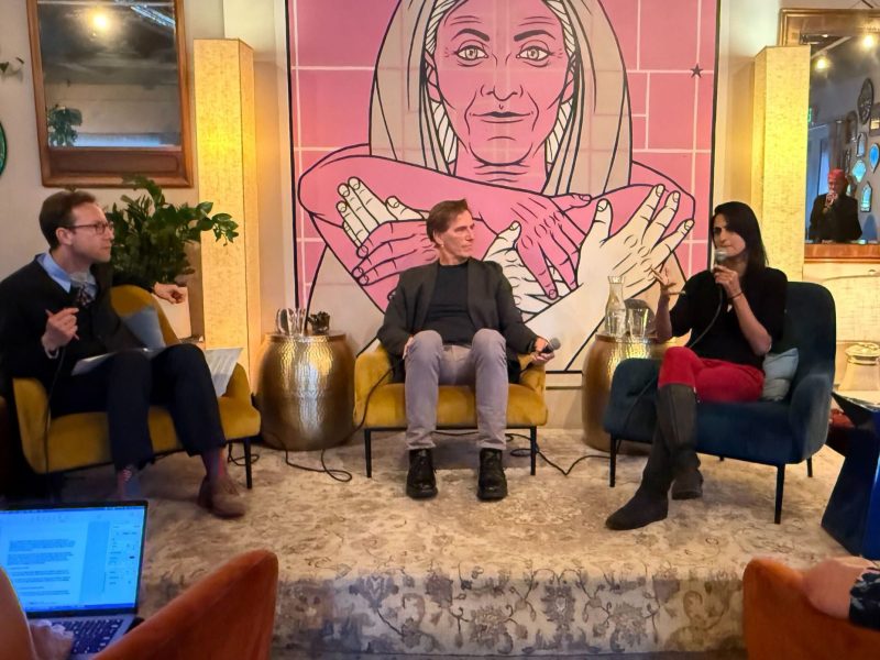 Three people sit and converse on a small stage in a cozy room, discussing harm reduction, with a large mural of a woman and the word "TOGETHER" behind them. Audience members are visible in the foreground.