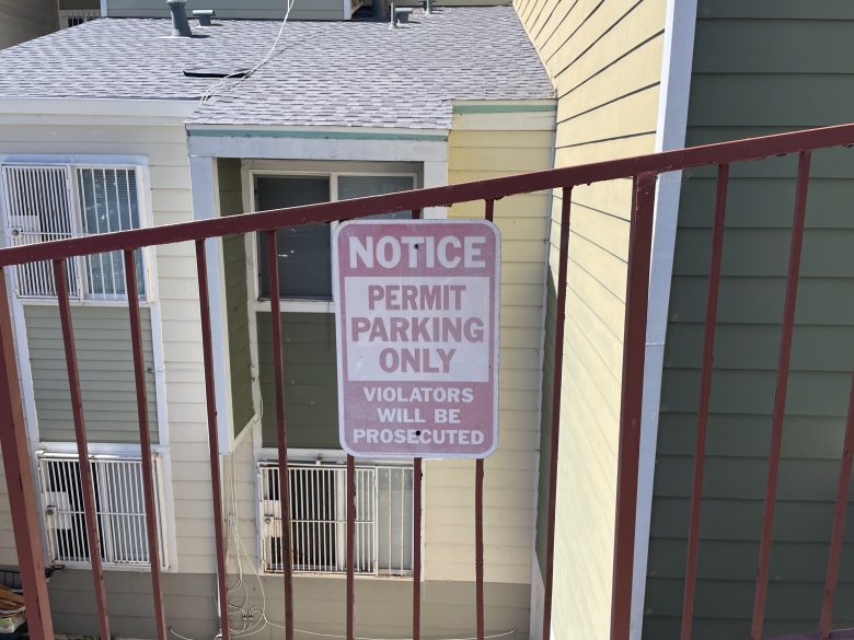 A sign on a metal railing reads, "NOTICE PERMIT PARKING ONLY VIOLATORS WILL BE PROSECUTED," with apartment buildings in the background.