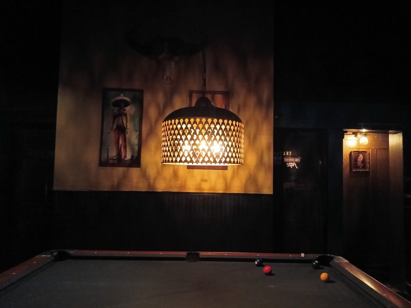 A dimly lit room with a pool table, three balls on the table, a hanging lamp, and framed pictures on the wall.