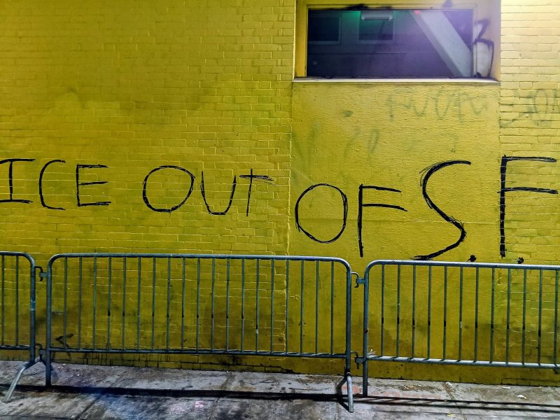 Yellow brick wall with black spray-painted graffiti reading "ICE OUT OF S.F." Metal barricades are placed in front of the wall, and a window is visible at the top right.