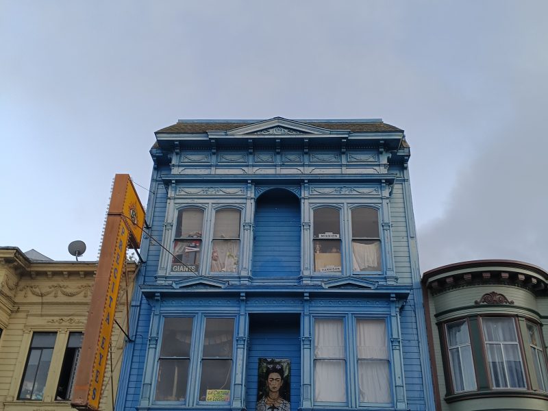 A blue three-story building with bay windows houses the Precita Eyes Mural Arts and Visitors Center, with a mural of a woman above the yellow entrance.