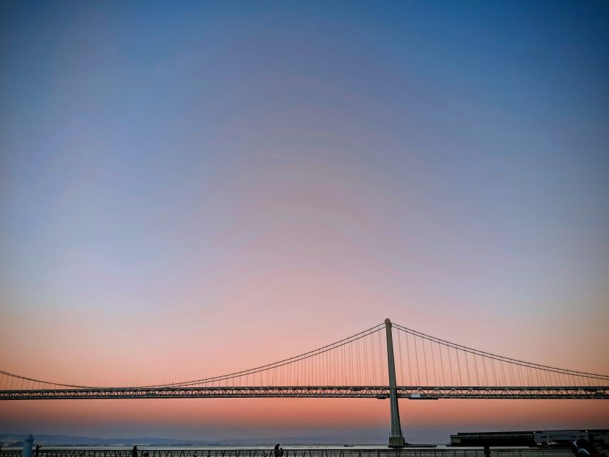 A suspension bridge stretches across the horizon beneath a clear sky at dusk, with soft pink and blue tones in the background.