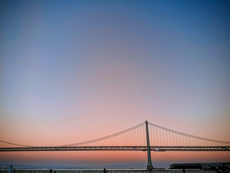A suspension bridge spans across calm water at sunset, with a clear gradient sky transitioning from pink near the horizon to blue above.