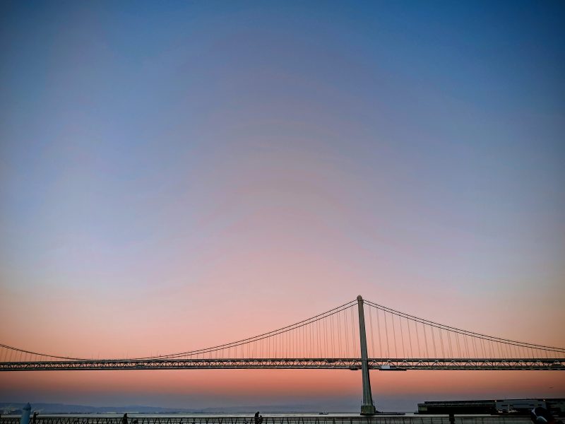 A suspension bridge spans across calm water at sunset, with a clear gradient sky transitioning from pink near the horizon to blue above.