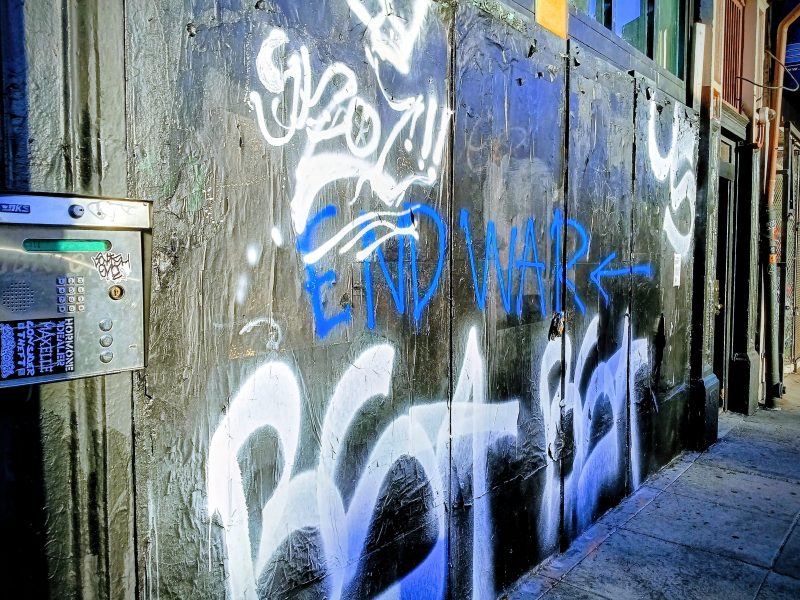 A black-painted wall with white and blue graffiti, including the words "END WAR" written in blue, next to a keypad entry system on a city sidewalk.