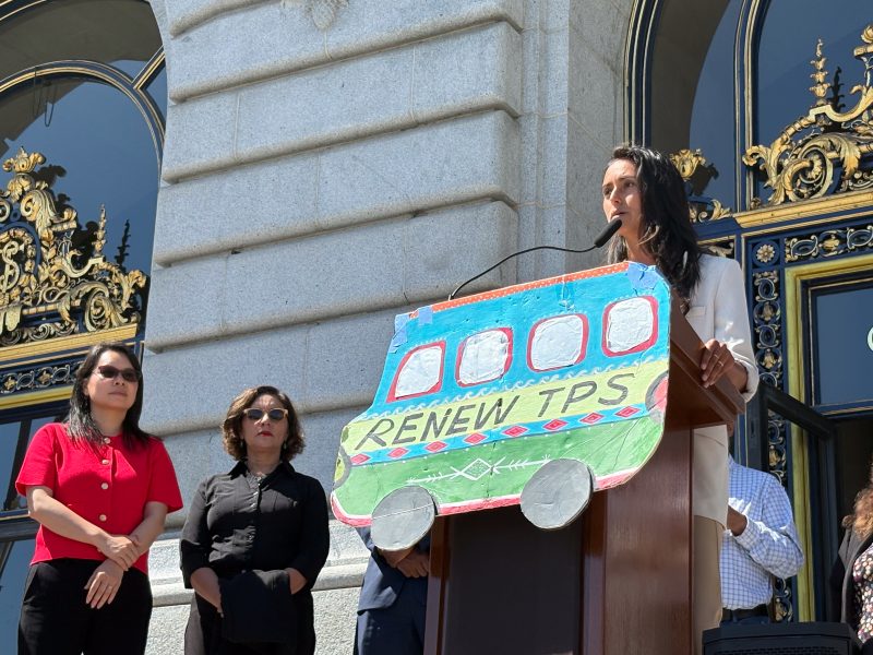 A woman speaks at a podium decorated with a sign reading "Renew TPS," while two other women stand nearby in front of a government building.