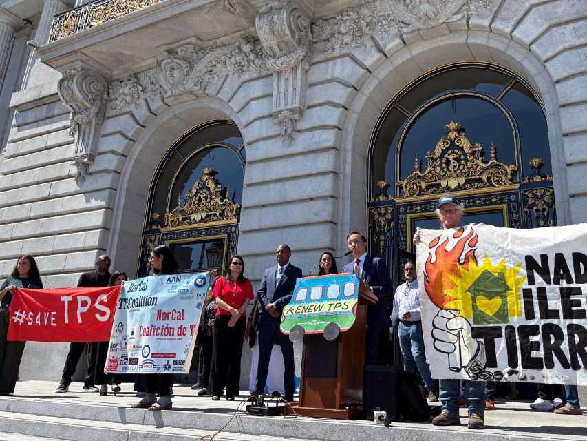 A group of people stand on steps in front of a building holding signs supporting TPS and climate action during a public rally or press event.