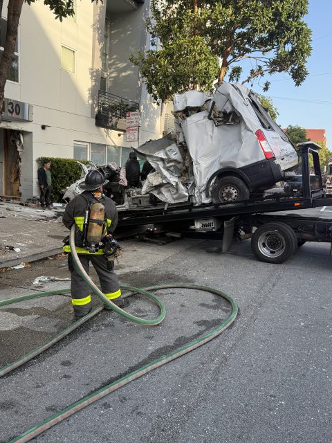 A firefighter observes as a severely crushed van is loaded onto a tow truck in front of a damaged building.