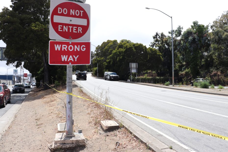 A "Do Not Enter" and "Wrong Way" sign stands beside a road blocked by yellow caution tape, with police cars parked nearby and trees lining the area.
