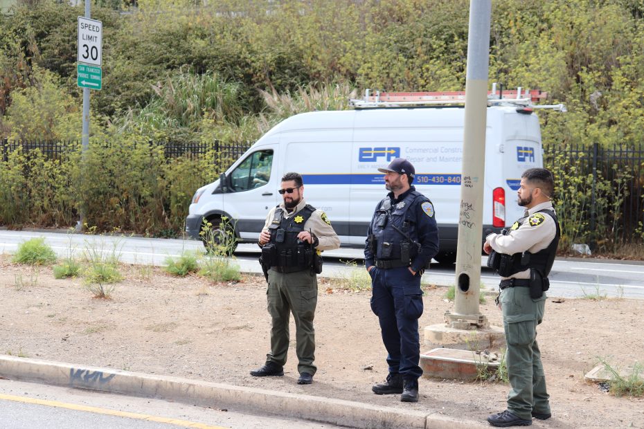 Three uniformed police officers stand on a sidewalk beside a street; a white commercial van is parked in the background near a speed limit sign.
