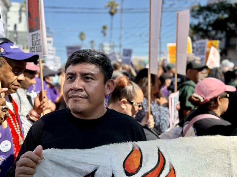 A man stands at the front of a crowd holding a banner during a daytime protest; other people in the background hold signs and banners.