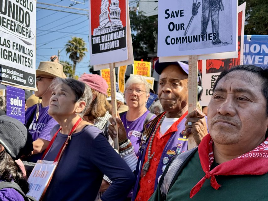 A diverse group of protesters hold signs with messages such as "Save Our Communities" and "The Billionaire Takeover" during a daytime demonstration.