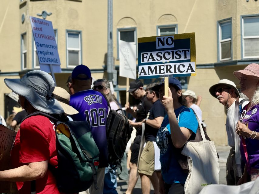 People participate in a protest; one person holds a sign reading "NO FASCIST AMERICA," while others carry various signs. Buildings are visible in the background.