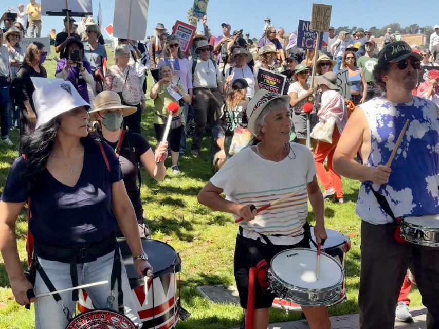 A group of people play drums at an outdoor protest or rally, while a large crowd stands behind them holding signs and wearing hats.