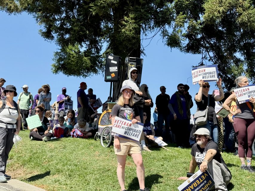 A group of people gather outdoors, some standing and some sitting on grass, holding signs that read "Veteran Care Not Corporate Welfare.