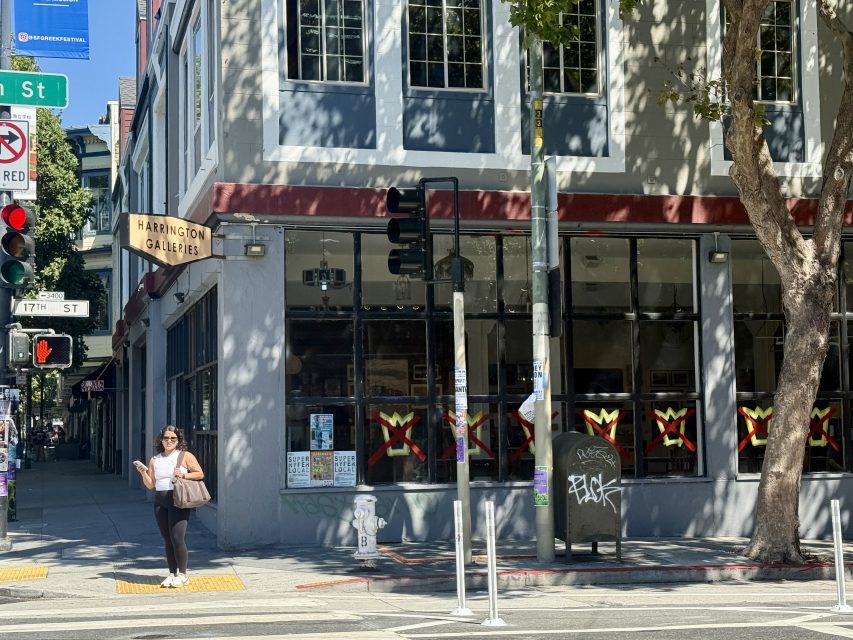A woman stands at a street corner near a building with a "Harrington Galleries" sign and large windows. There are traffic lights, a tree, and a graffiti-marked utility box nearby.