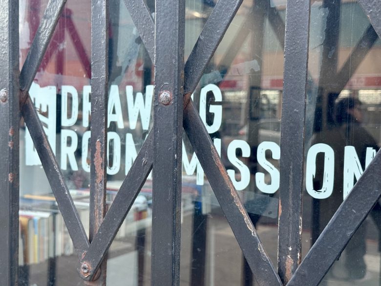 A black metal gate with an X-shaped pattern partially obscures a glass window with white painted text and shelves of books visible behind it.