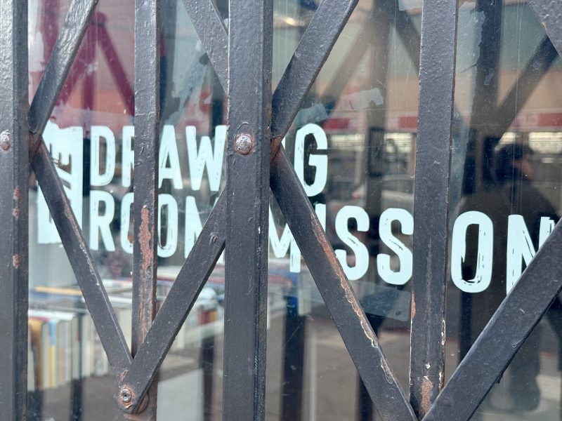 A black metal gate with an X-shaped pattern partially obscures a glass window with white painted text and shelves of books visible behind it.