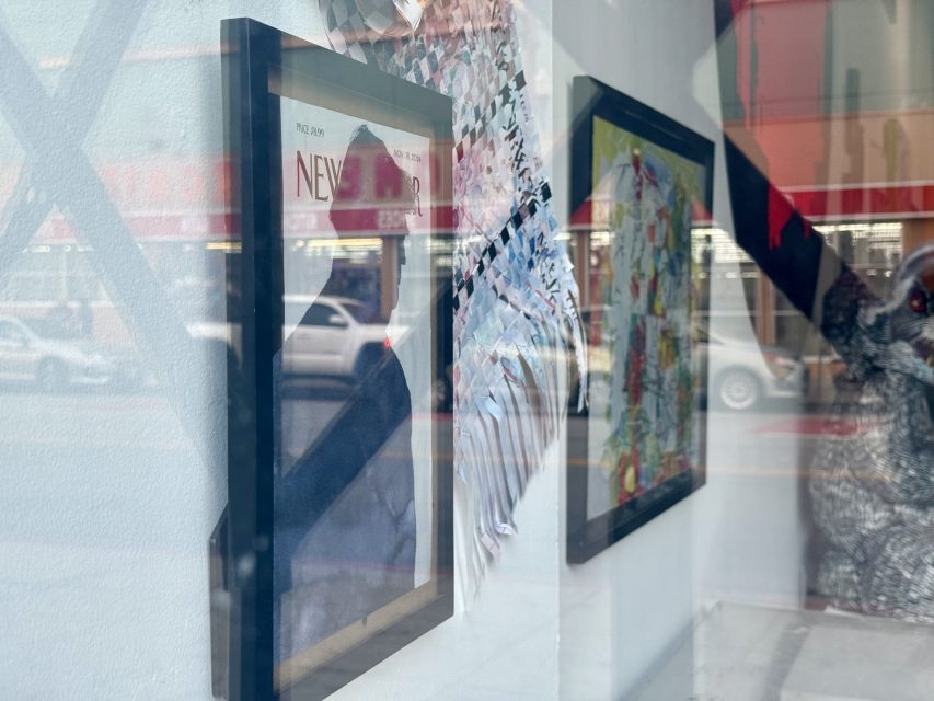 Framed artwork displayed on a white wall behind glass, with reflections of a street, cars, and buildings visible in the window.
