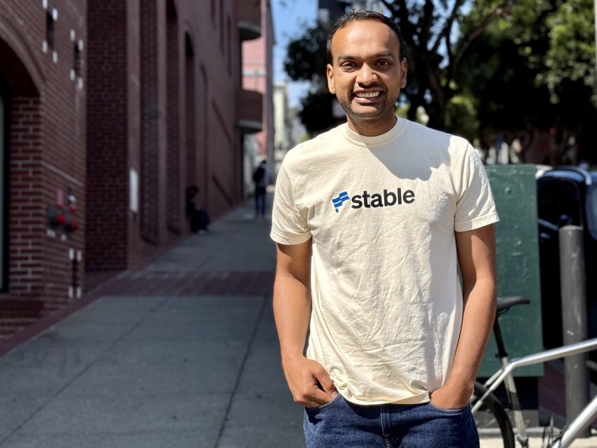 A person stands on a sunny city sidewalk, wearing a cream-colored "Stable" t-shirt and blue jeans, with a bicycle and buildings in the background.