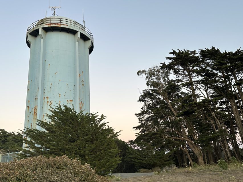 A tall, cylindrical, weathered light blue water tower stands next to tall trees under a clear sky.
