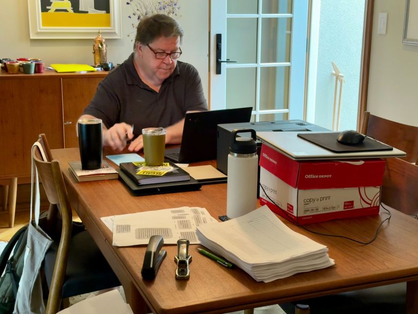 A man works at a dining table cluttered with papers, folders, drinks, office supplies, and a laptop, trying to recall important details in the sunlit home office setting.
