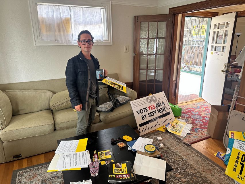 Person standing in a living room holding a yellow recall campaign sign, surrounded by election materials and paperwork spread across the coffee table.