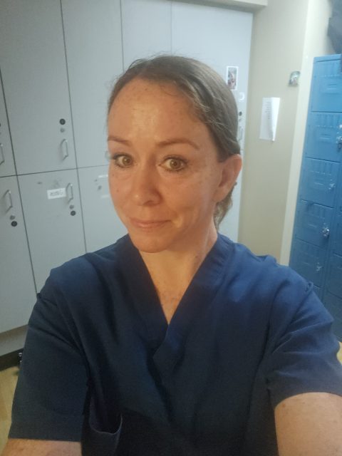 Person wearing blue scrubs taking a selfie in a room with lockers and cabinets in the background.