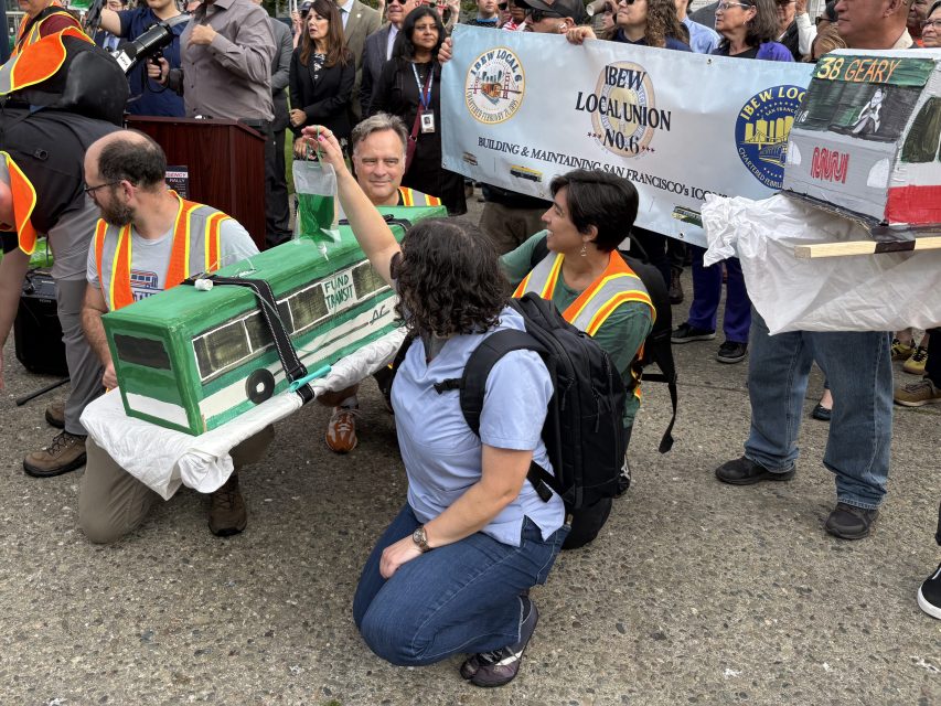A group of people wearing safety vests pose with a model bus at a crowded outdoor protest, holding signs related to public transit and labor unions.