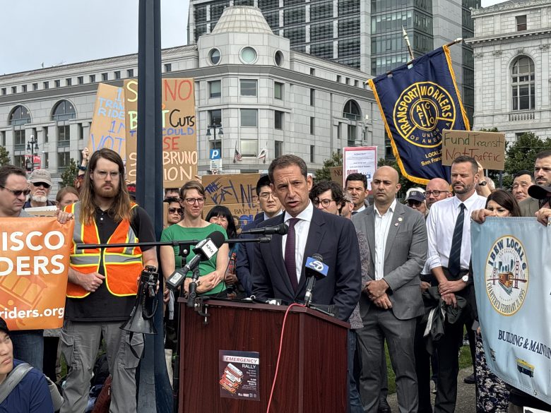 A man in a suit speaks at a podium during an outdoor rally, surrounded by people holding signs about transit and workers' rights.