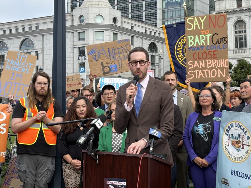 A man in a suit speaks at a podium during a protest. People behind him hold signs opposing BART service cuts and supporting public transit funding.