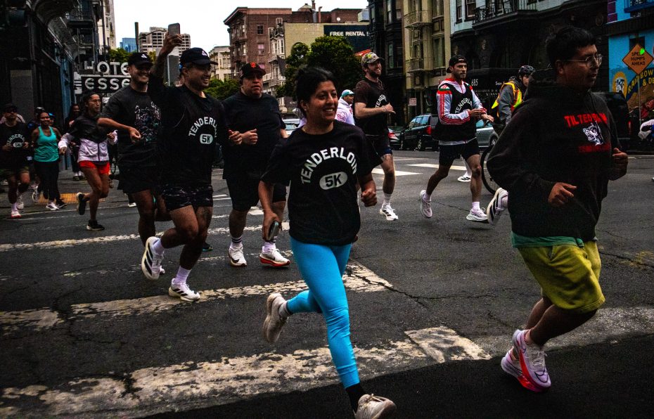 A group of people run across a city street during a 5K race, wearing athletic clothing and race t-shirts.