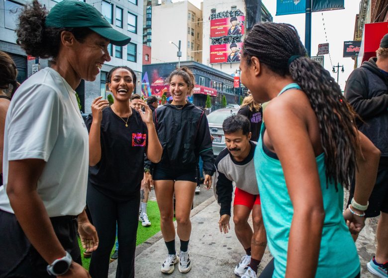 A group of people smile and laugh together on a city sidewalk, some wearing athletic clothing, with buildings and billboards in the background.