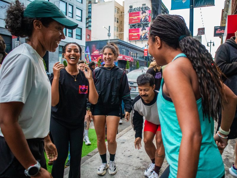 A group of people smile and laugh together on a city sidewalk, some wearing athletic clothing, with buildings and billboards in the background.