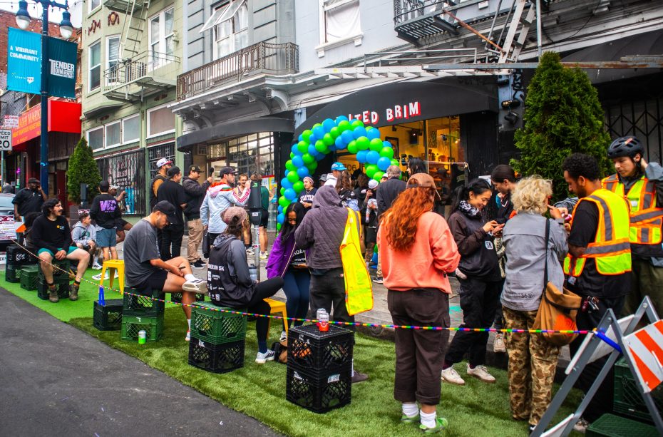 A group of people gather on a city street with tents, balloons, and barriers, some standing and some seated on crates, in front of a store with a "Smoked Brim" sign.