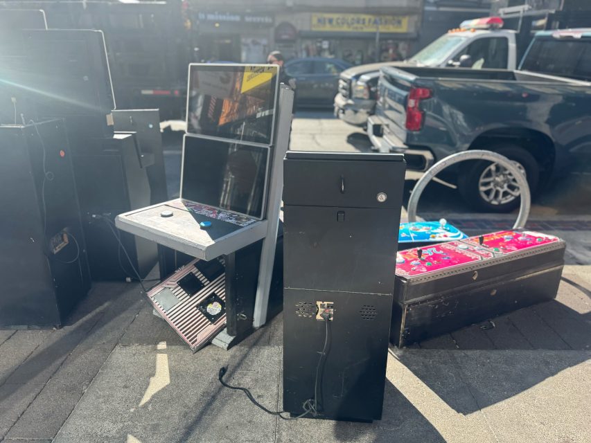 Several dismantled arcade machines and parts are placed on a city sidewalk, with vehicles and storefronts visible in the background.