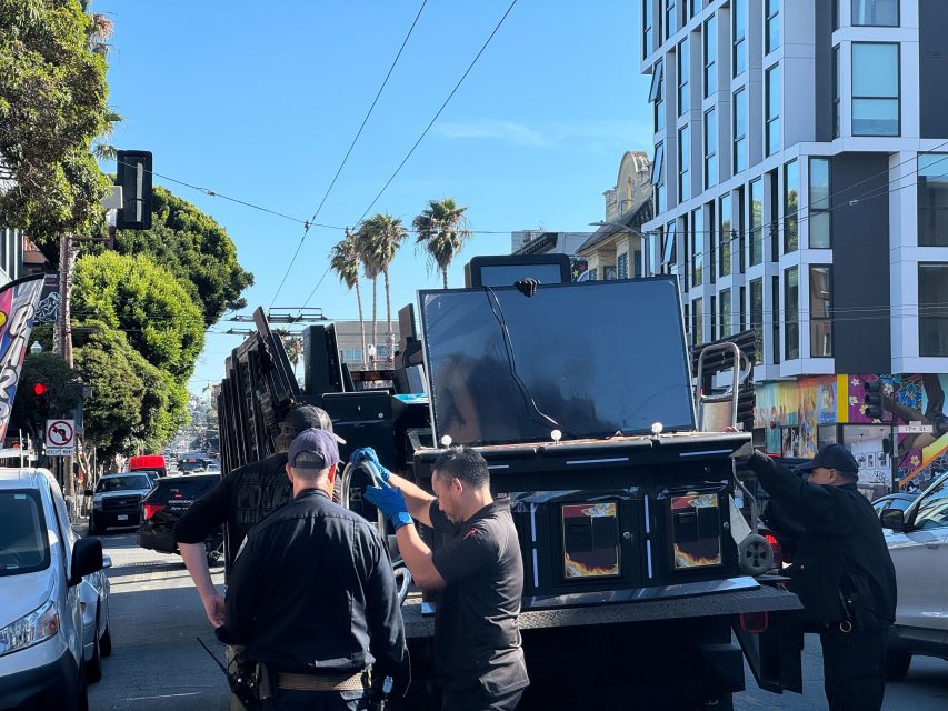 Workers transport a large flat-screen TV on a cart along a city street, surrounded by cars and modern buildings under a clear sky.