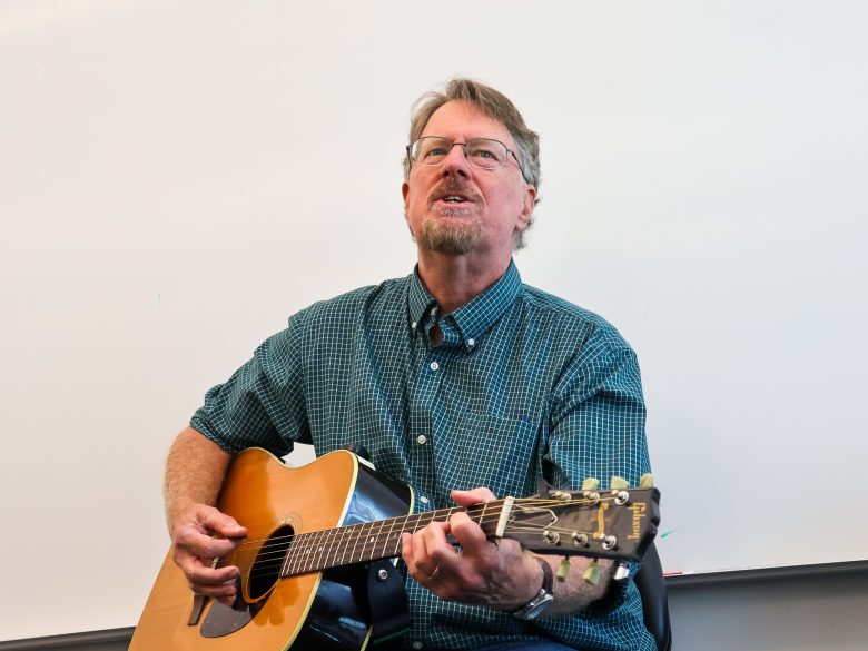 A man wearing glasses and a green checkered shirt plays an acoustic guitar while seated in front of a whiteboard.
