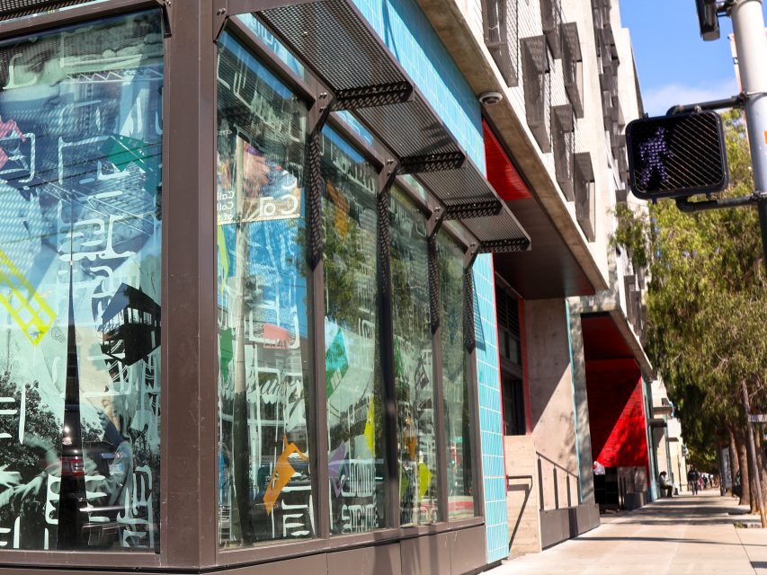 Glass storefront with abstract art on the windows on a city street corner, modern building facades, and a crosswalk signal visible.